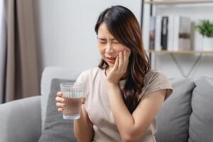 le femme avec une mal aux dents est en portant sa joue dans douleur et une verre de l'eau. photo