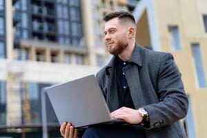 portrait de une Beau Jeune homme travail sur portable en plein air. mode homme est assis sur banc dans ville centre. flou Contexte. photo