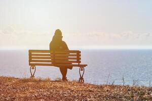 fille célibataire dans une veste noire et un chapeau assis sur un banc à la falaise au bord de la mer endroit calme et paisible photo