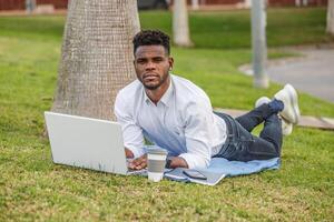 un Afro-américain homme est vu mensonge sur le herbe, en utilisant une portable ordinateur. photo
