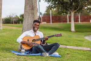 une souriant homme séance dans le vert herbe, grattage une guitare dans le chaud Soleil. photo