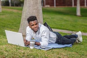 un africain américain homme travail sur le sien portable mensonge sur le sol dans le parc. photo