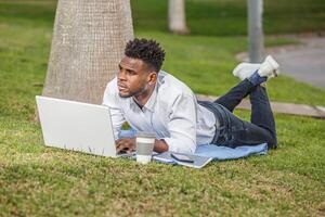 une homme mensonge sur le herbe tandis que en utilisant le sien portable ordinateur à travail photo