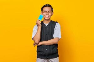 souriant jeune homme asiatique tenant une carte de crédit à la main et regardant la caméra sur fond jaune photo