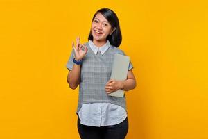 Portrait d'une femme asiatique joyeuse tenant un ordinateur portable et faisant un geste ok isolé sur fond jaune photo