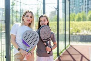 positif mère et fille permanent sur tribunal avec padel raquettes pendant Pause et à la recherche à caméra. photo