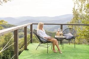 une Jeune femme, pigiste est travail sur une portable à distance sur une balcon dans le ouvert Frais air près le montagnes dans chaud ensoleillé temps. photo