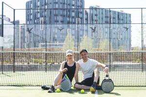 portrait de positif Jeune femme et adulte homme permanent sur padel tennis rechercher, en portant raquette et balle, souriant photo