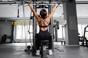 musclé athlétique femelle bodybuilder dans noir costume tiré sur des sports simulateur dans salle de sport. retour muscles sont très tendu. photo