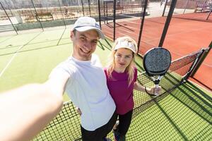 Jeune couple de tennis joueurs portant une tenue de sport en portant une raquette sur leur épaules et une Balle dans une main permanent Extérieur sur tennis tribunal à de bonne heure Matin photo