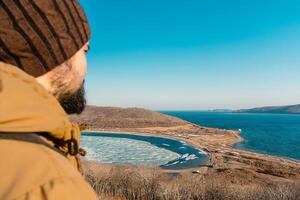 une homme dans une chapeau et veste regards à le mer horizon et le collines avec jauni herbe. une homme est randonnée le long de le vallonné mer rivages. russky île, vladivostok, Russie. photo