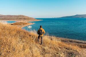 une homme dans une chapeau et veste regards à le mer horizon et le collines avec jauni herbe. une homme est randonnée le long de le vallonné mer rivages. russky île, vladivostok, Russie. photo