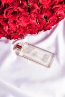 une transparent élégant parfum bouteille avec une délicat fragrance mensonges sur une blanc satin Contexte. rouge des roses bouquet. verticale Haut voir. Vide allonger photo