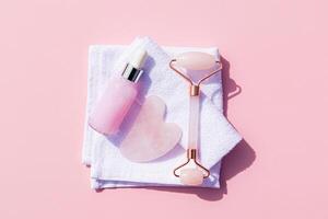 rouleau masseur, gua sha grattoir, Naturel massage pétrole dans une bouteille avec une compte-gouttes sur une blanc serviette. rose Contexte. santé et beauté concept photo