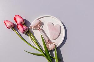 Rose quartz rouleau masseur et grattoir sur une rond céramique plateau avec vivre tulipes. Haut voir. gris Contexte. Naturel se soucier. photo