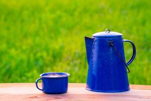 bouilloire, bleu émail, et café des tasses sur un vieux en bois sol, flou Contexte de riz des champs à lever du soleil. photo