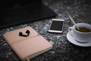 sur le noir marbre table sont une ordinateur portable, sans fil écouteurs une cellule téléphone, une bloc-notes, une tasse de thé photo