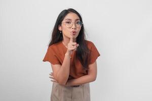 Jeune asiatique femme portant marron chemise et monocle à la recherche à caméra tandis que en mettant une doigt dans de face de lèvres faire des gestes rester silencieux ou rester calme, isolé par blanc Contexte. photo