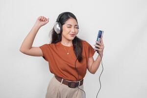 excité Jeune asiatique femme dans marron chemise élevage serré poing geste tandis que écoute à la musique de téléphone intelligent en utilisant sans fil casque de musique. photo