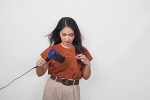 attrayant asiatique femme dans marron chemise sentiment dérangé tandis que en utilisant sèche-cheveux en essayant à faire coiffure, isolé plus de blanc Contexte. photo