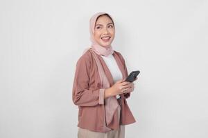 souriant Jeune asiatique musulman femme portant hijab et en portant une téléphone intelligent isolé par blanc Contexte. photo