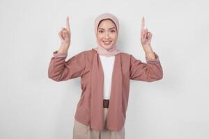 excité asiatique musulman femme portant hijab montrer du doigt à le copie espace à l'envers son, isolé par blanc Contexte. photo