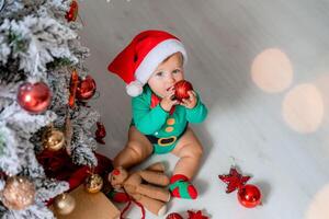 mignonne bébé dans une vert le maillot de corps et une Père Noël chapeau décore le Noël arbre avec coloré des ballons photo