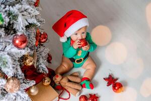 mignonne bébé dans une vert le maillot de corps et une Père Noël chapeau décore le Noël arbre avec coloré des ballons photo