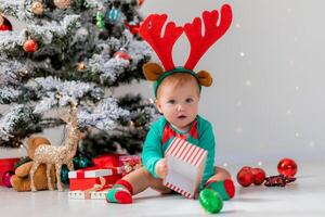 bébé dans une vert le maillot de corps avec rouge cerf cornes sur le sien tête est en jouant suivant à le Noël arbre photo