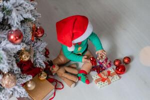 mignonne bébé dans une vert le maillot de corps et une Père Noël chapeau décore le Noël arbre avec coloré des ballons photo