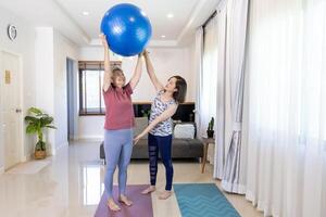 Sénior asiatique femme est exercice à Accueil en utilisant yoga Balle pour coeur corps et abdomen muscle bâtiment avec sa entraîneur fille ajustement le pose et encourager pour aîné en bonne santé photo