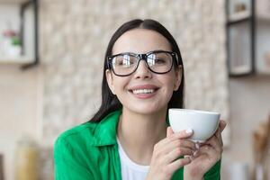 fermer photo portrait de une Jeune magnifique femme dans lunettes, en portant et en buvant une chaud boisson dans le matin, souriant et à la recherche en dehors le fenêtre