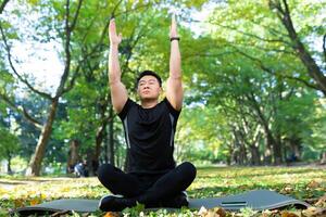 asiatique homme dans le parc sur une des sports tapis est assis dans le lotus position, sportif effectue respiration des exercices sur une ensoleillé jour, homme dans une des sports costume repose après physique et aptitude des exercices photo