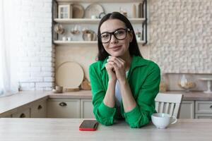 Jeune magnifique femme dans des lunettes et une vert décontractée chemise, à Accueil dans le cuisine, regards à le webcam, appel écoute à le interlocuteur, vue de le webcam photo