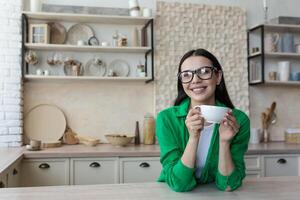 Jeune magnifique femme dans des lunettes et une vert décontractée chemise, à Accueil dans le cuisine, regards à le webcam, appel écoute à le interlocuteur, vue de le webcam photo