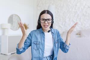 magnifique femme à la recherche à la toile caméra, nerveux et argumenter avec interlocuteur, en utilisant casque pour appel, travail à Accueil séance sur canapé, en ligne conférence et étude photo