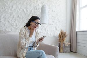 une Jeune magnifique femme à Accueil est séance sur le canapé dans le vivant chambre, souriant joyeusement, en portant une téléphone intelligent dans sa mains, recevoir le joyeux nouvelles de confirmation de admission à Université photo