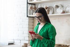 une Jeune magnifique femme est permanent à Accueil dans le cuisine près le fenêtre avec une téléphone dans sa mains. ordres nourriture maison, fait du achats dans un en ligne magasin. photo