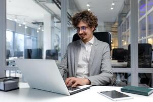 de bonne humeur et réussi homme d'affaire à l'intérieur Bureau travail avec ordinateur portable, barbu homme souriant et dactylographie sur portable clavier, programmeur codage Logiciel dans moderne chambre. photo