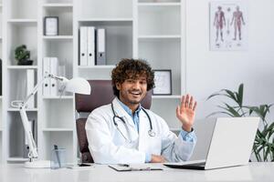 souriant Jeune hispanique Masculin médecin étudiant, interne, assistant séance dans Bureau à ordinateur portable, en étudiant, Faire pratique, qui passe examen. salue avec le sien main, présente lui-même à le caméra. photo