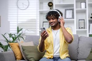 Jeune souriant hispanique homme à Accueil écoute à la musique en utilisant en ligne app sur téléphone, homme souriant seul à Accueil séance sur canapé dans vivant pièce en train de regarder diffusion . photo