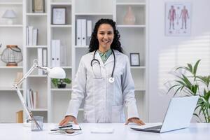 portrait de une souriant Jeune réussi musulman femelle médecin permanent à une bureau dans un Bureau dans une uniforme et à la recherche à le caméra. photo