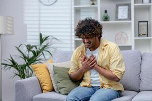 malade homme ayant sévère poitrine douleur et cœur attaque, hispanique Jeune homme séance seul à Accueil dans vivant chambre, en portant mains à poitrine. photo