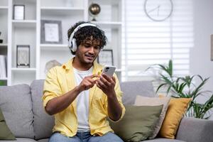 Jeune souriant hispanique homme à Accueil écoute à la musique en utilisant en ligne app sur téléphone, homme souriant seul à Accueil séance sur canapé dans vivant pièce en train de regarder diffusion . photo