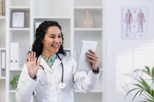Jeune hispanique femelle médecin permanent dans clinique Bureau parlant sur appel avec patient, consultant en ligne, salutation et agitant à tablette caméra. photo