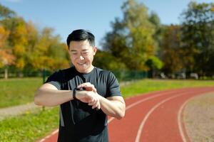 un asiatique homme, un athlète permanent sur une fonctionnement Piste dans une stade, à la recherche et vérification le temps sur une intelligent montre. il sourires, satisfait avec le résultat de le courir, le marathon, rupture le enregistrer. photo