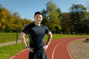 portrait de une souriant et sur de soi asiatique Masculin athlète permanent dans de face de le caméra dans une stade avec le sien bras à le sien côtés. photo