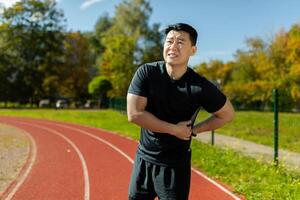 asiatique sportif a sévère douleur dans retour et abdominal muscles, homme faire pipi formation dans stade sur ensoleillé journée masser côté de dos, athlète sur le jogging tiré muscle. photo