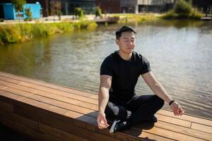 asiatique Jeune Masculin athlète, entraîneur, praticien est assis par le Lac sur une banc dans le lotus position. il se ferme le sien yeux, médite, Est-ce que yoga, repose. photo