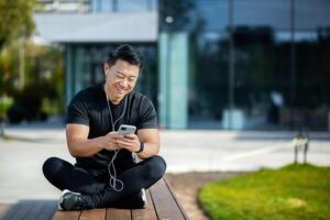 asiatique Jeune Masculin sportif séance sur banc dans lotus position près Lac dans parc portant écouteurs et écoute à musique, podcast, parlant sur téléphone. repos. photo